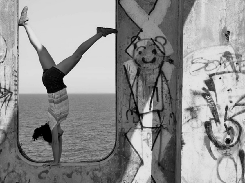 Person doing handstand in graffiti-framed window by ocean.